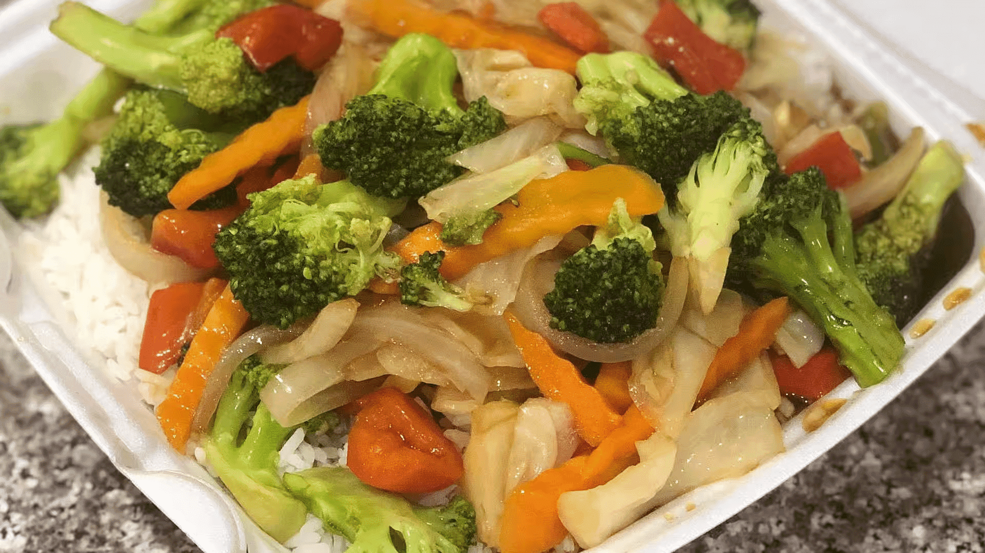 Stir-fried vegetables with broccoli, bell peppers, and onions served over white rice in a take-out container.