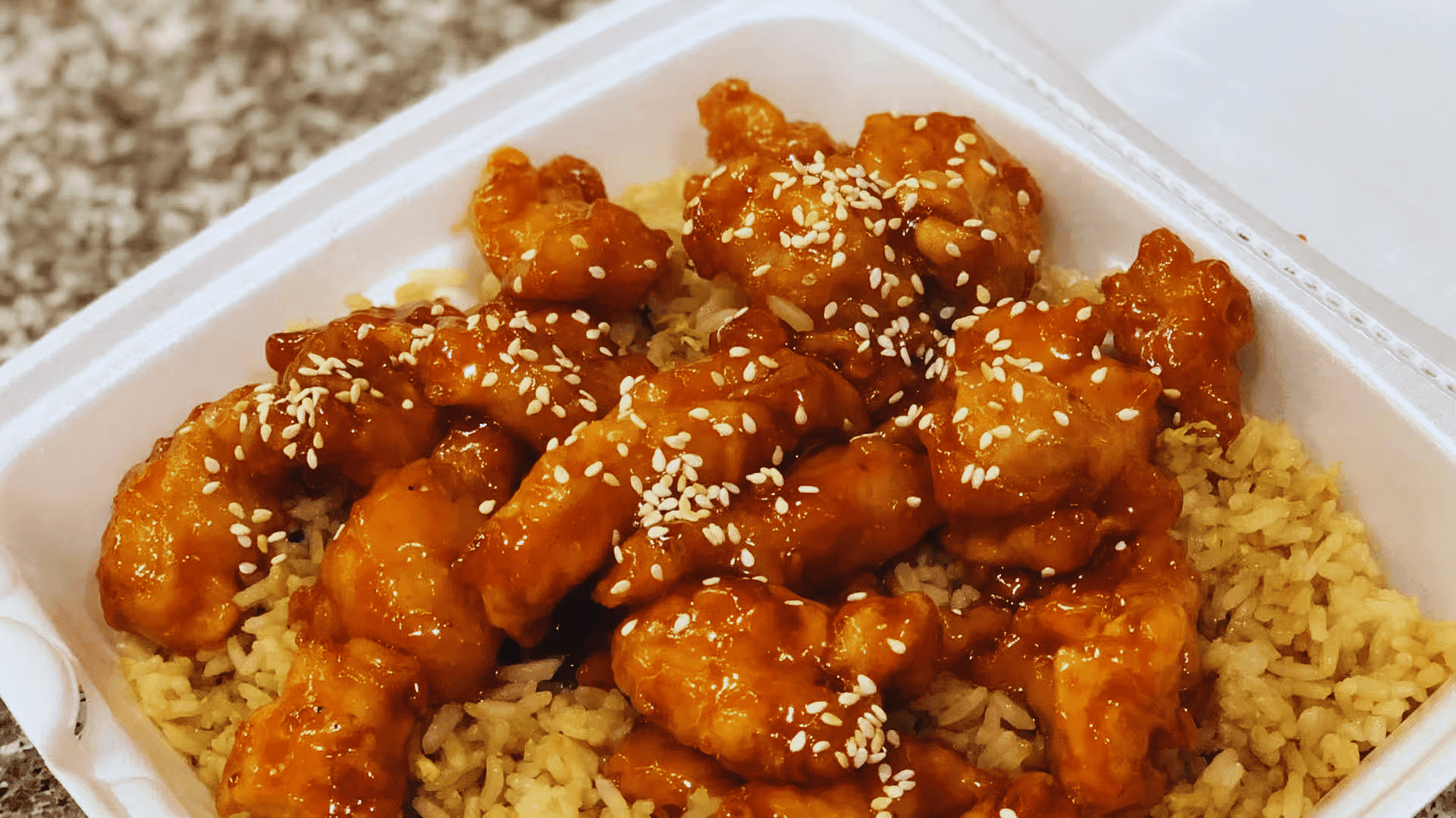 Sesame chicken served over fried rice in a white takeout container.