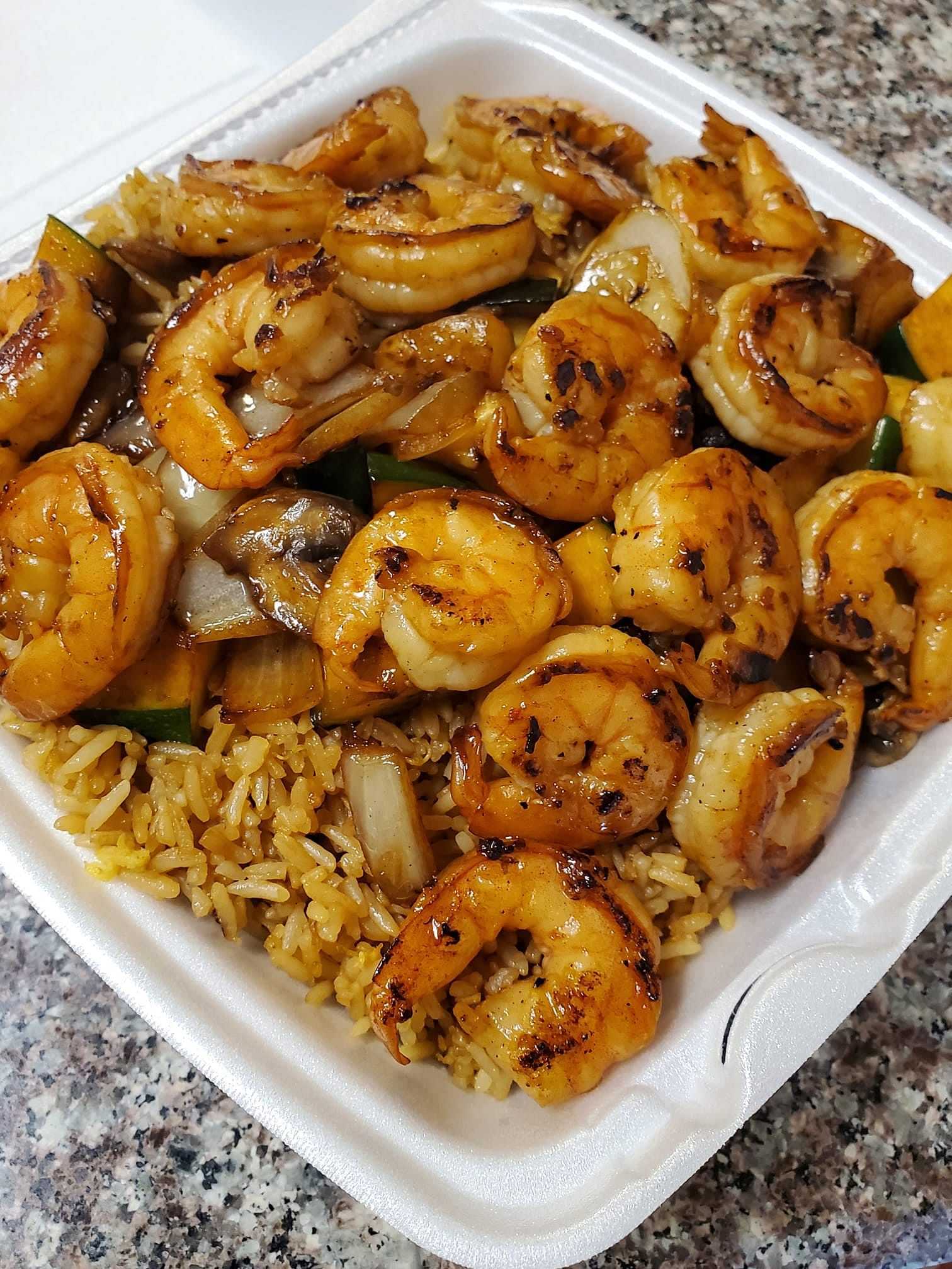 Grilled shrimp with vegetables served on fried rice in a takeout container.