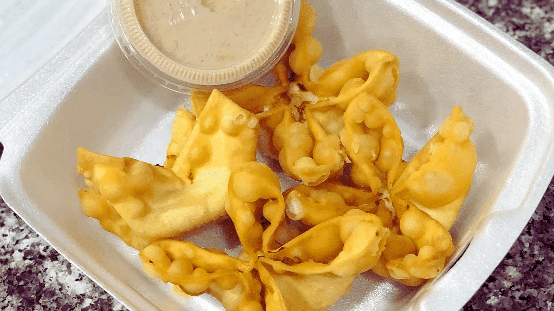 Crispy wontons with dipping sauce in a takeout container placed on a granite countertop.