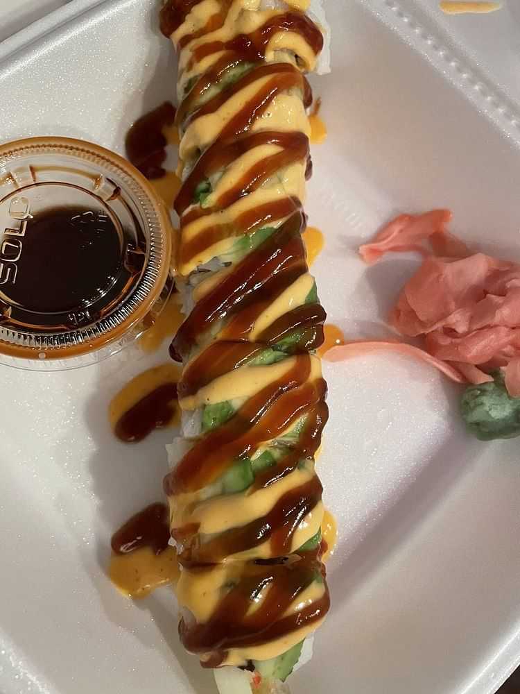 Sushi roll drizzled with sauces next to a container of soy sauce, ginger, and wasabi in a takeout box.